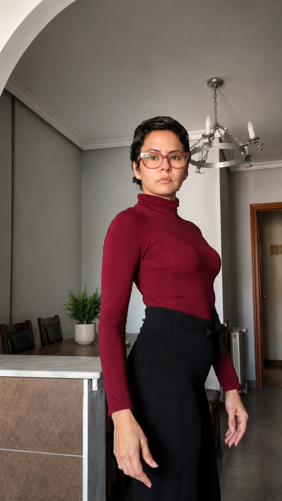 Mujer con cabello corto y gafas, usando cuello alto borgoña y pantalón negro, de pie en un espacio interior moderno y minimalista.